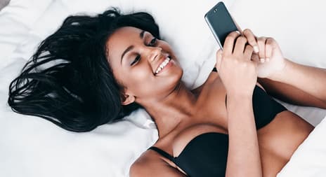 Dame i sengen som holder en smartphone og smiler.